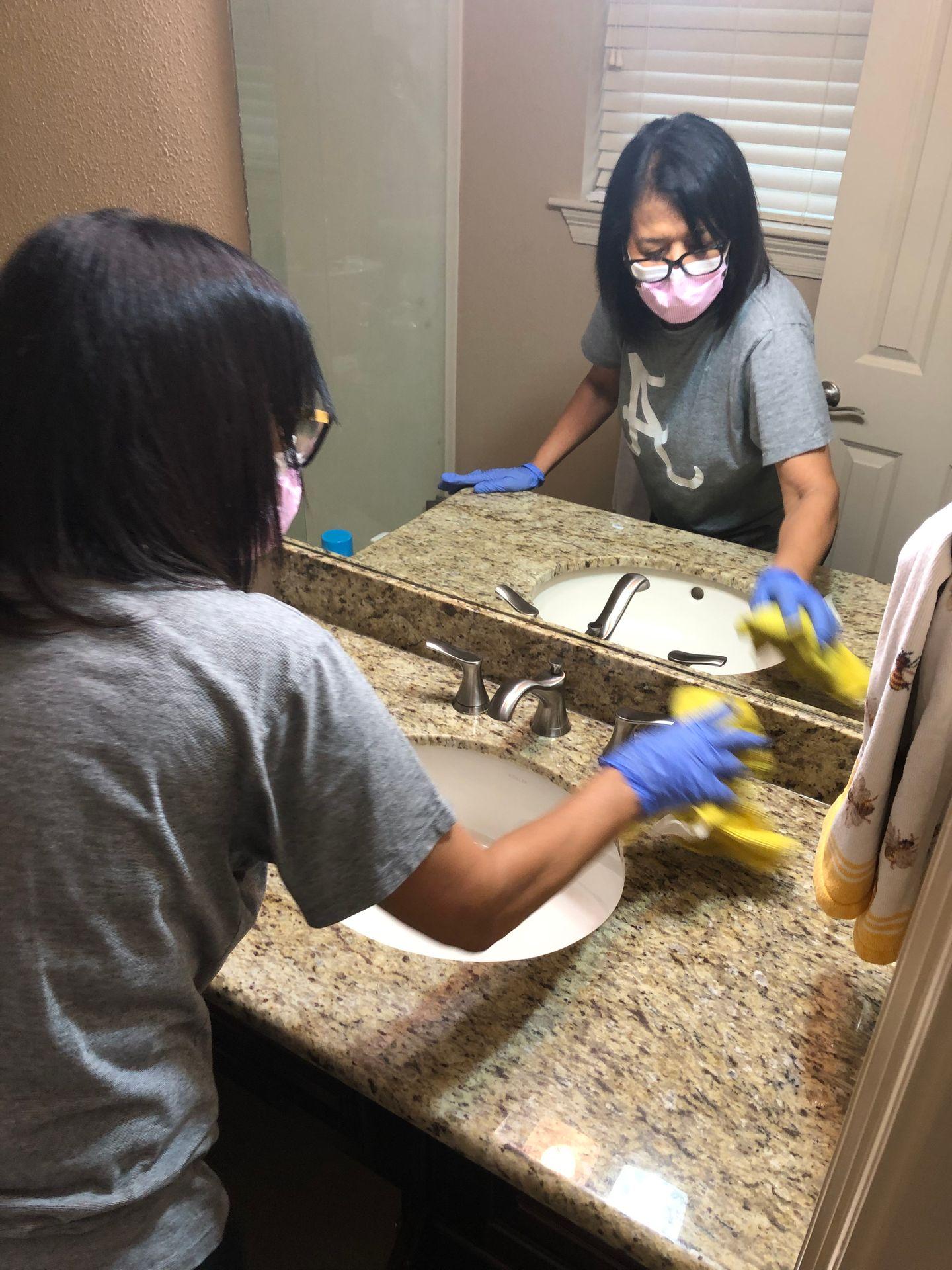 Person wearing gloves and a mask cleaning a bathroom sink with a cloth.